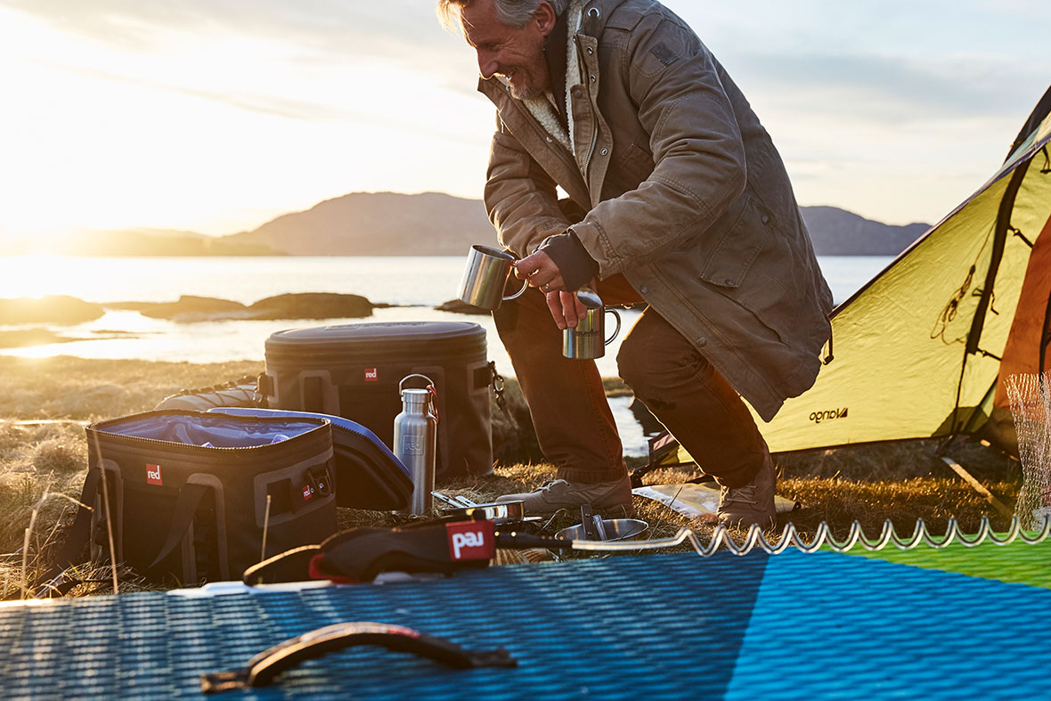 Man Camping On A Beach With 2 Red Original Cool Bags, A Drink Bottle & A Red Paddle Board