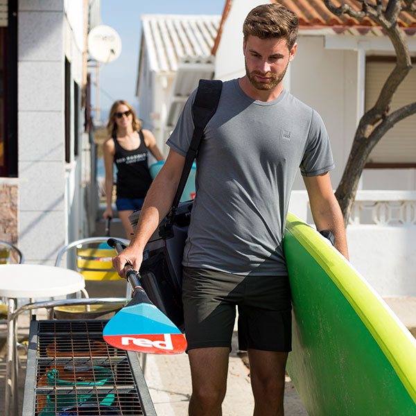 Man walks carrying paddle board wearing Performanc T shirt.
